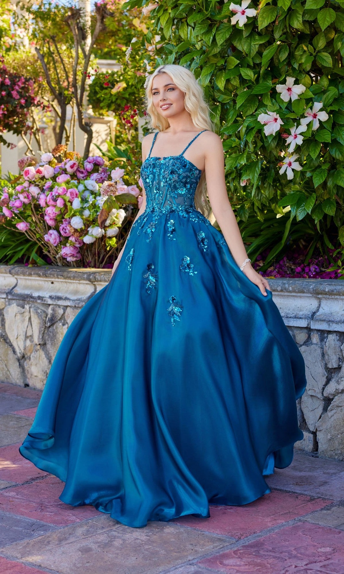 A woman in the teal Juliet JT2617U long prom dress, featuring a lace-up back and embellished A-line design, stands outdoors on a stone patio amid flowers and greenery.